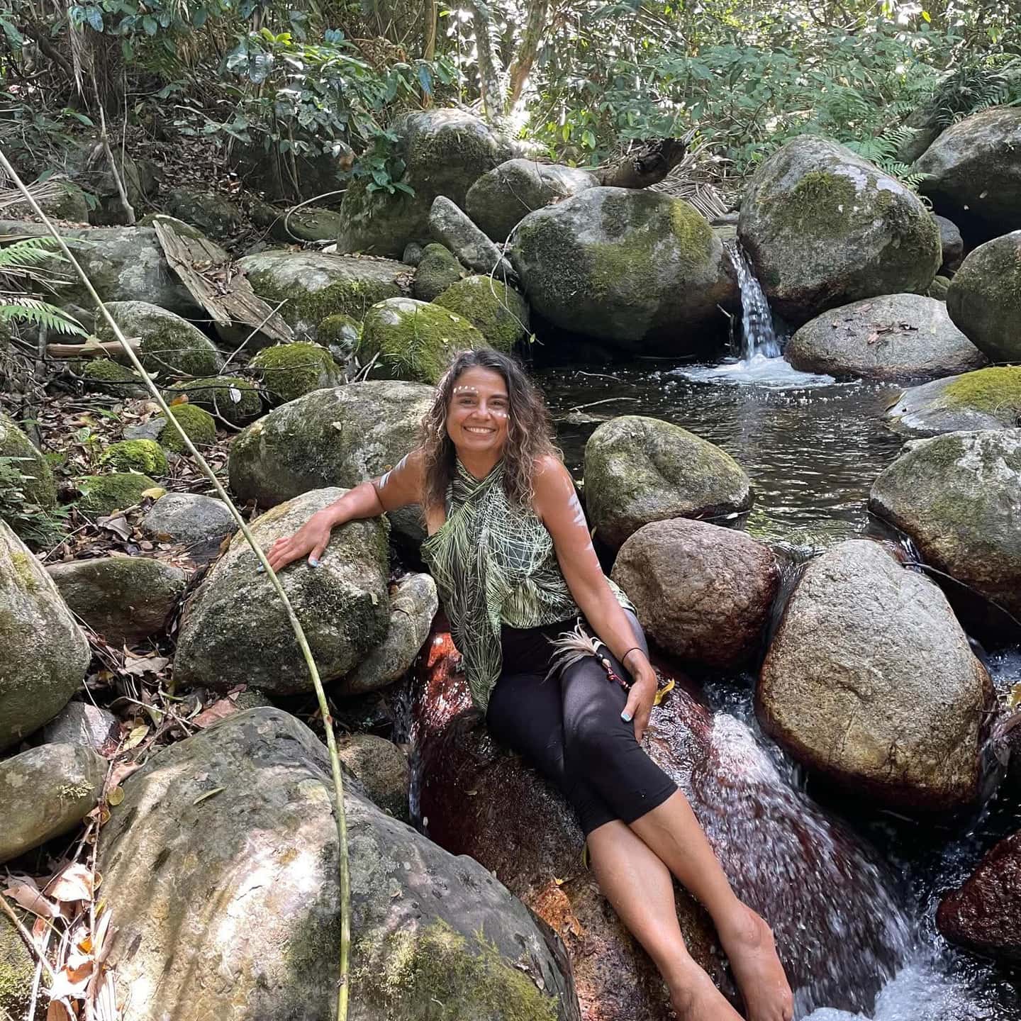 Julie smiling beside a small rainforest waterfall among moss-covered rocks on Country.