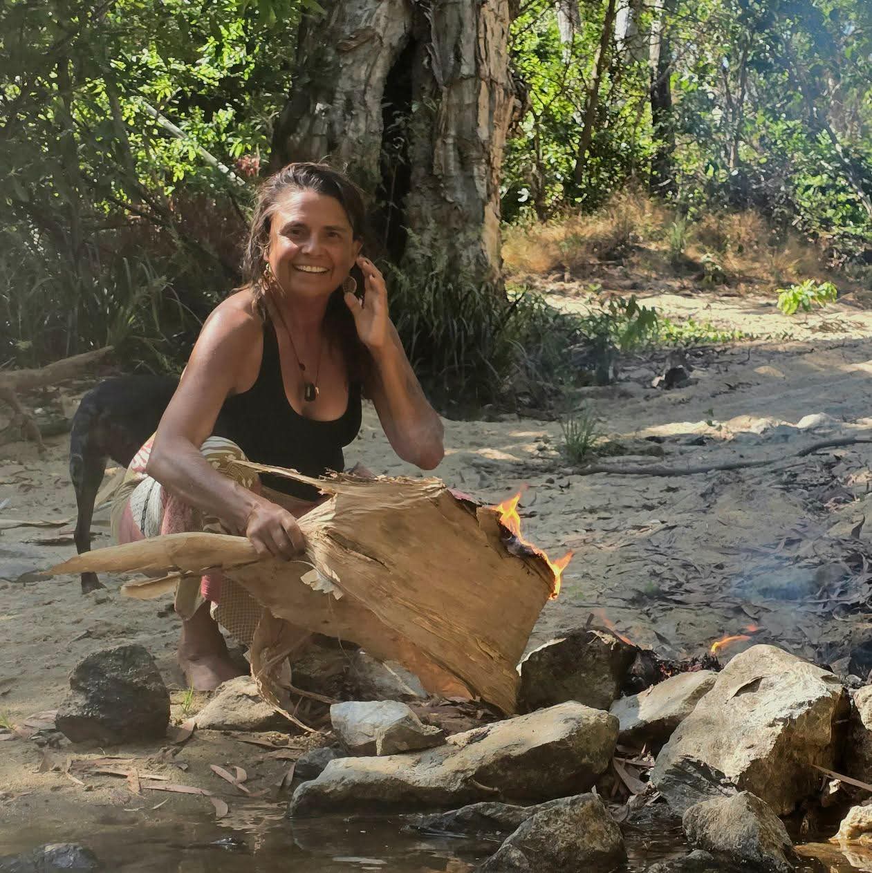 Julie smiling while tending a small fire beside the creek on Country.