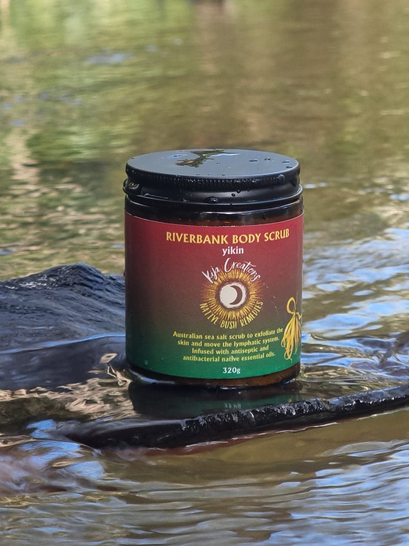 A Riverbank Body Scrub jar resting beside creek water.